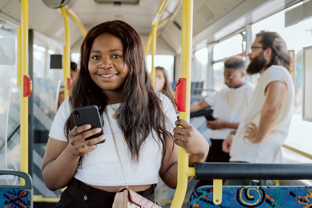 A dark-skinned woman rides in a public transport bus holding on to a barrier, a girl buys an electronic ticket on her phone through an app, other passengers in the vehicle in the background