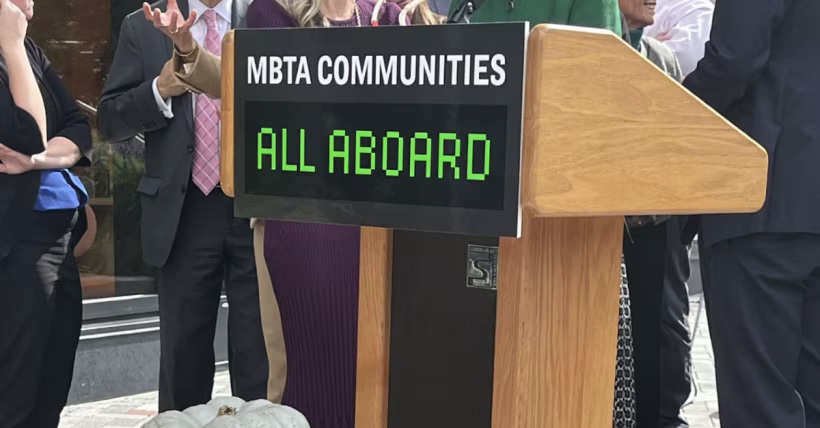 A front view of a podium with a sign saying “MBTA COMMUNITIES ALL ABOARD”