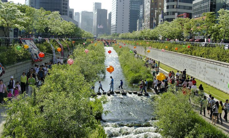 Cheonggyecheon in Seoul, South Korea. The former overpass is now a river surrounded by vegetation with people walking on adjacent sidewalks.
