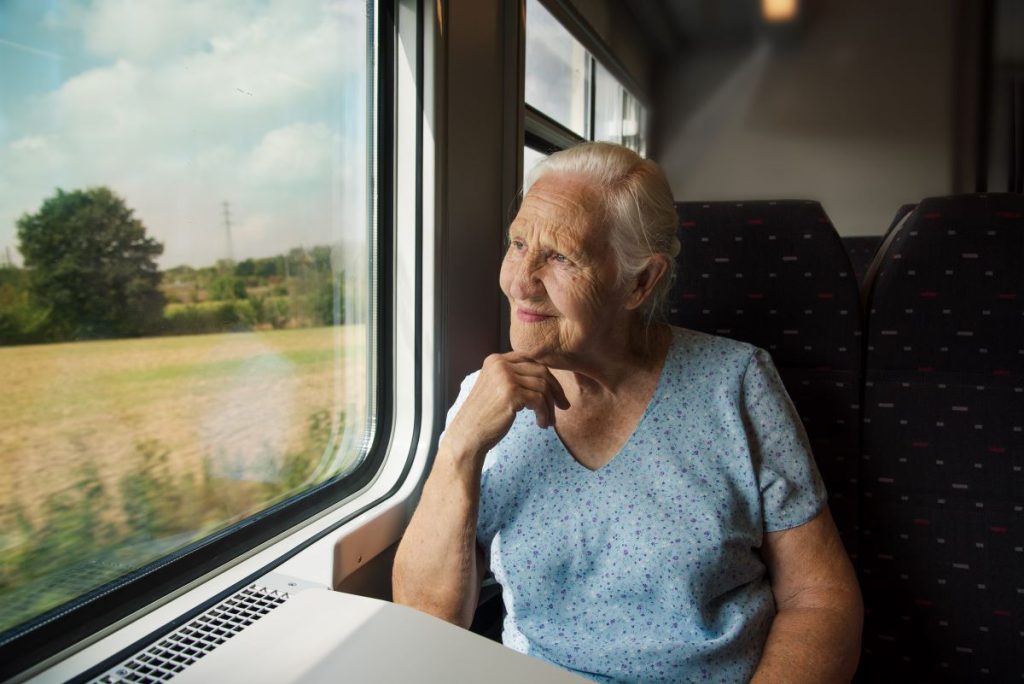 An elderly woman sitting on a train and smiling as she looks out the window.