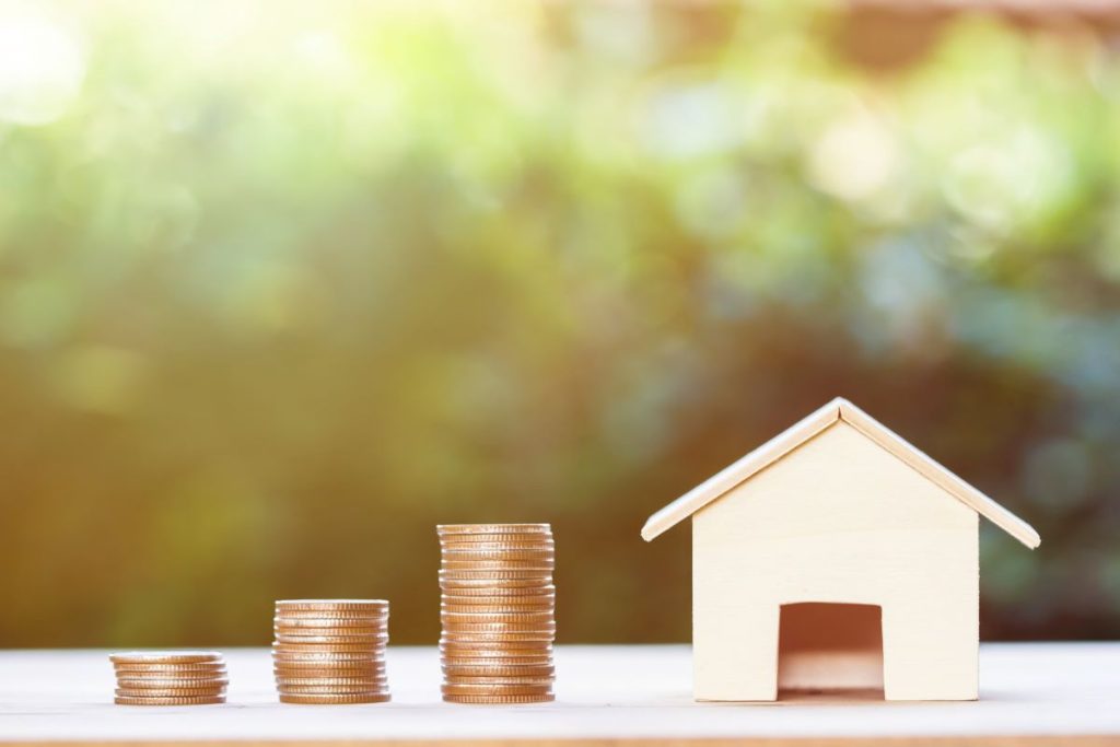A miniature paper house positioned next to a stack of coins. 