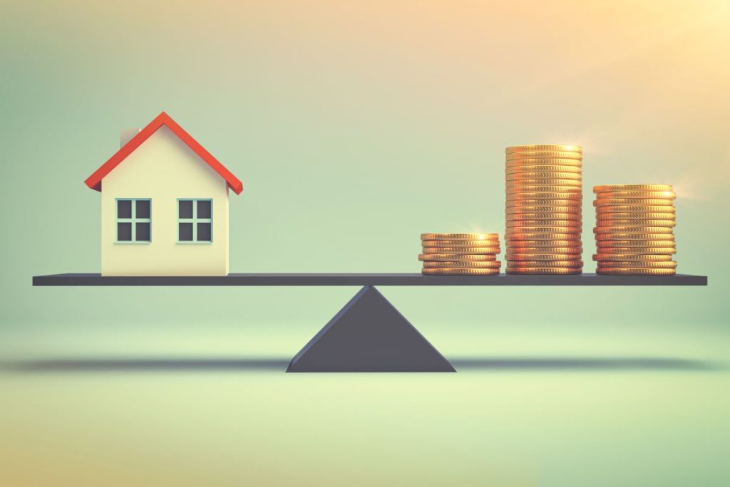 A miniature house balancing on one side of scales with a pile of coins on the other.