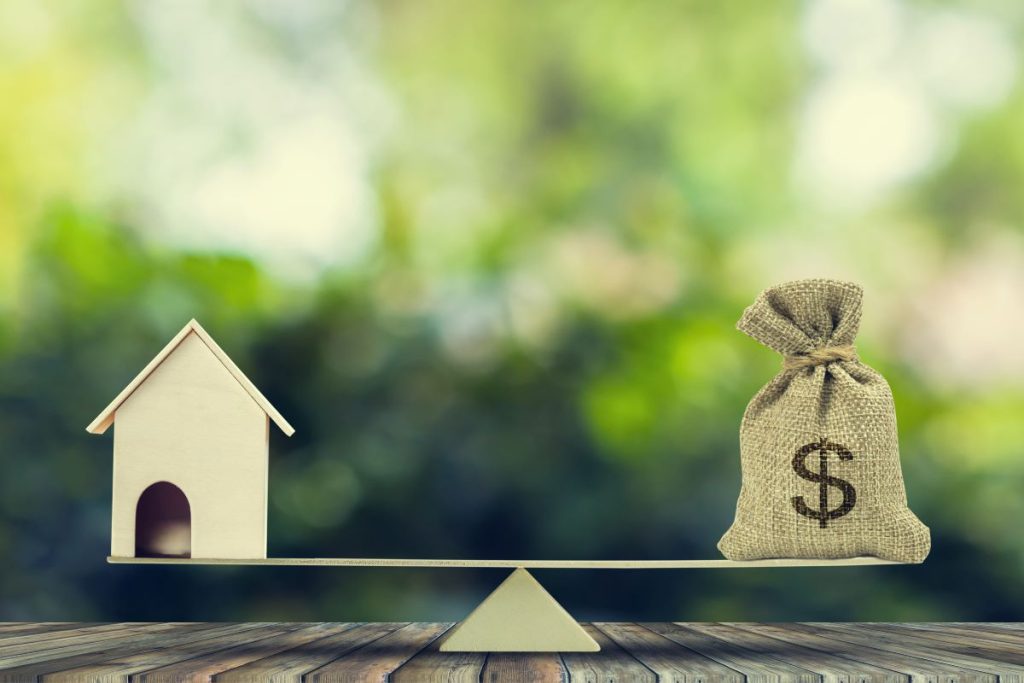 A miniature wooden house balancing on a beam with a small bag of money on the other.