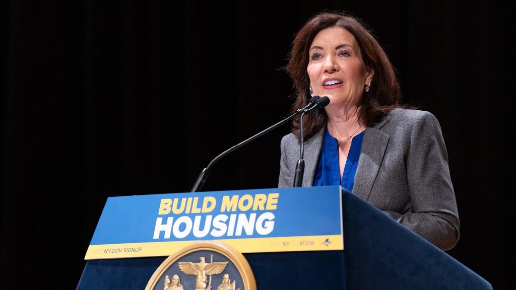 Kathy Hochul standing at a podium that says "Build More Housing"